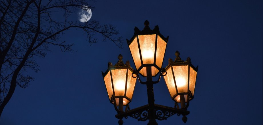 Eine Straßenlaterne leuchtet, im Hintergrund sieht man den Mond. 